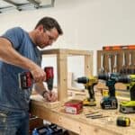 Man working with cordless drill and tools in a woodworking workshop.