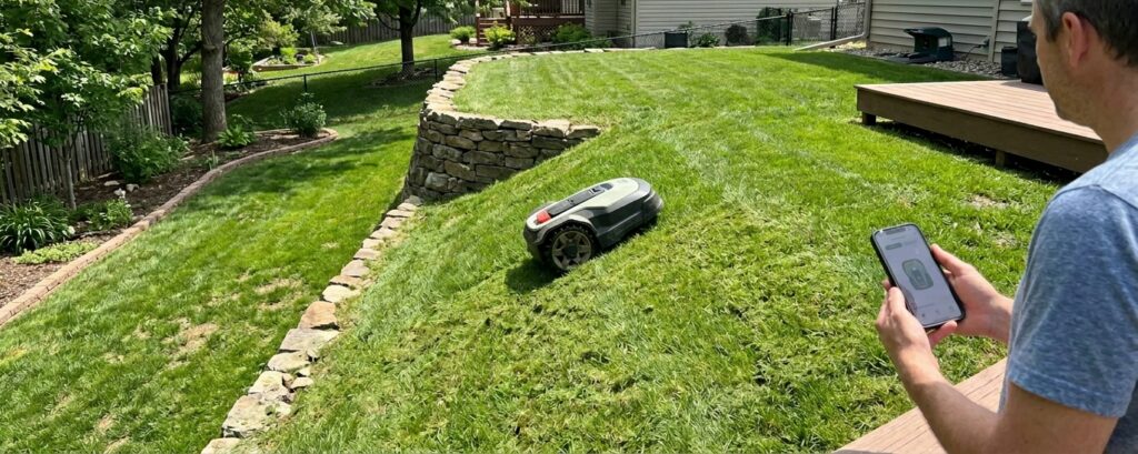 Robot mower navigating steep lawn on a hillside with a person controlling via smartphone.