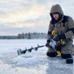 Man kneeling on snow with ice auger in winter landscape.
