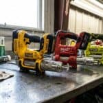 Multiple cordless grease guns from DeWalt, Milwaukee, RYOBI on workbench for maintenance tasks.
